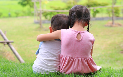 Older sister hugs little brother by the neck, shoulders sitting on green grass field.