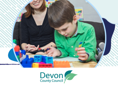 A young boy playing with colourful building blocks whilst an adult looks on.