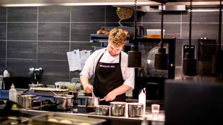 A man with red hair is in a restaurant kitchen. He is stirring something in a saucepan with a whisk. The saucepan is on a hob. The man is wearing chef's whites and a black apron with the word restaurant on it. There are several other saucepans on the counter, as well as ingredient containers and sauce bottles.