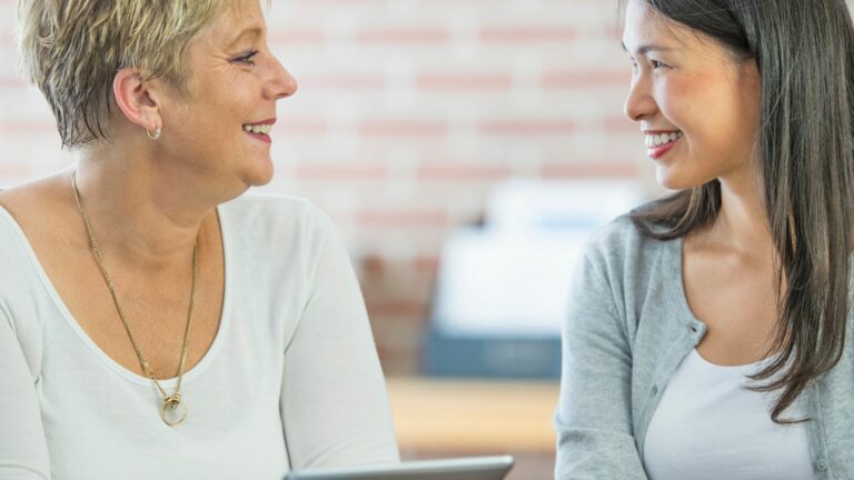 A middle aged white woman with short cropped hair is sat facing a young Asian woman. They are both smiling, and are in conversation. The white woman is holding a tablet computer and is sharing information with her colleague. The white woman is wearing a very ight blue long sleeve top, and a gold necklace. The Asian lady is wearing a grey cardigan.