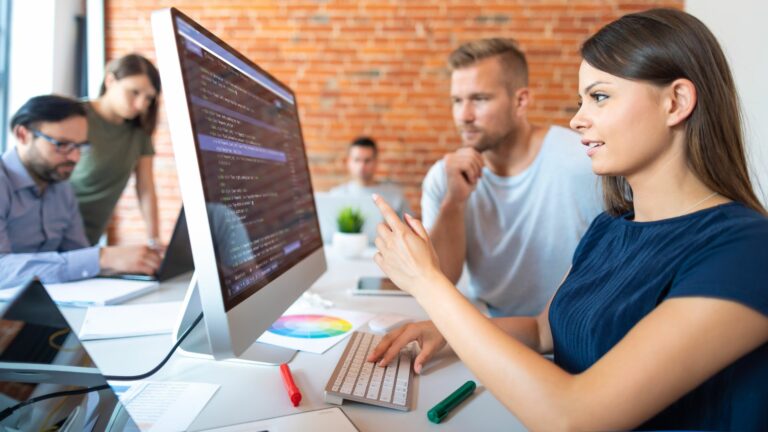 A white woman with long brown hair is pointing at a computer screen with code on it. A white man sat next to here is also looking at the screen, she is teaching him about what is on the screen. They are sat in an office and other colleagues are also working on laptops.