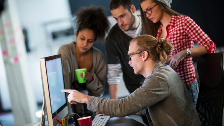 A white middle aged man who has long brown hair in a top knot, and big glasses is working at a computer he is pointing at a screen. There are three other people, a black woman holding a bright green mug, a white man with a beard and black short sleeve t-shirt over a white long sleeve top, and a white woman with black rimmed glasses and a red check shirt. They are looking quizzically at the screen. They are being taught by the older man.