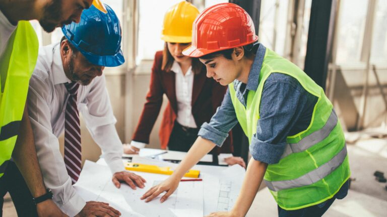 Four people are stood on a building site, they have plans on a table as well as spirit levels and pencils. They are in conversation. There is a woman wearing a high vis tabard, and a red hard hat who is leading the conversations. There is a man in a shirt and tie with a blue helment on, there is a woman in a yellow helmet and a red blazer with a white shirt. There is a man with a yellow helment and a high vis tabard over a white t-shirt.
