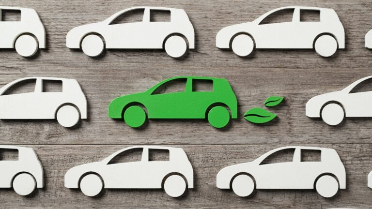 Nine paper cut out cars are displayed on a wooden background. eight are white, one is green and has green leaves coming out of the exhaust.