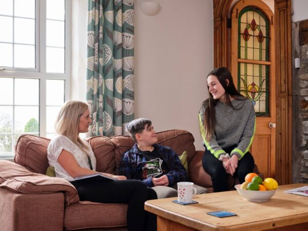 Two young people sat on a sofa with a social worker smiling.